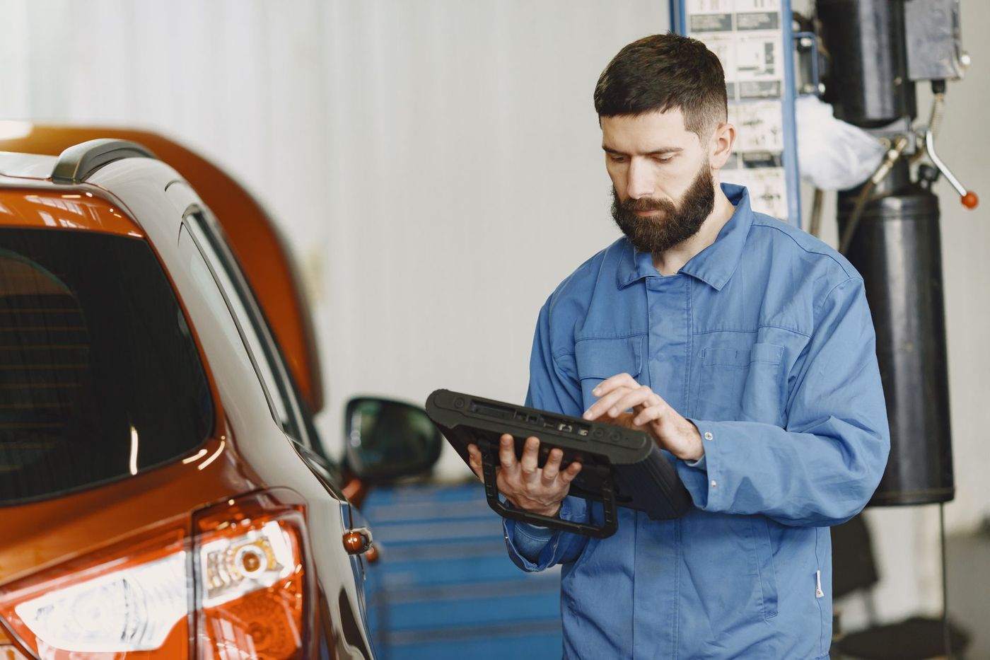 A technician in plain blue coveralls reading a handheld automotive diagnostic tool beside a car