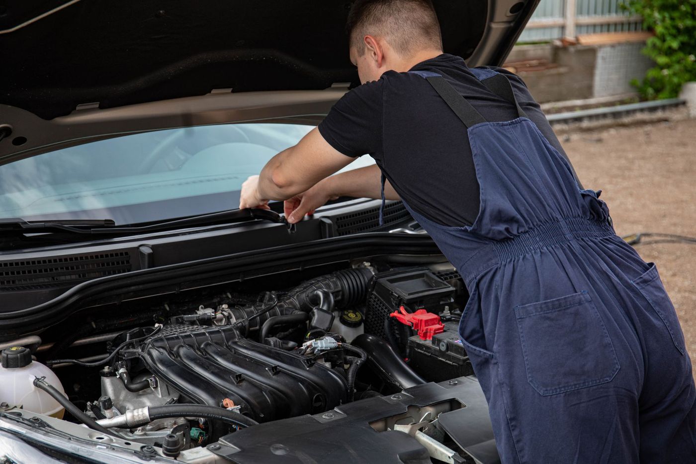 A mechanic in navy overalls leaning into an open car hood outdoors on gravel